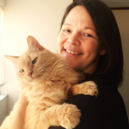 A smiling woman with medium length dark hair holding a large light orange cat with enormous paws.