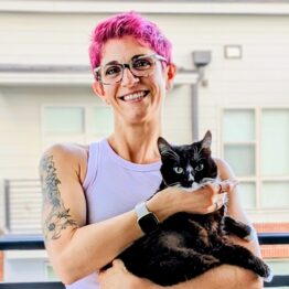 A smiling woman with short pink hair and glasses holding a black and white cat with an intense look on its face.